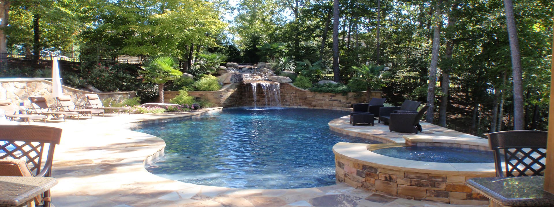 A backyard pool with a stone waterfall and attached spa, surrounded by a flagstone patio and trees