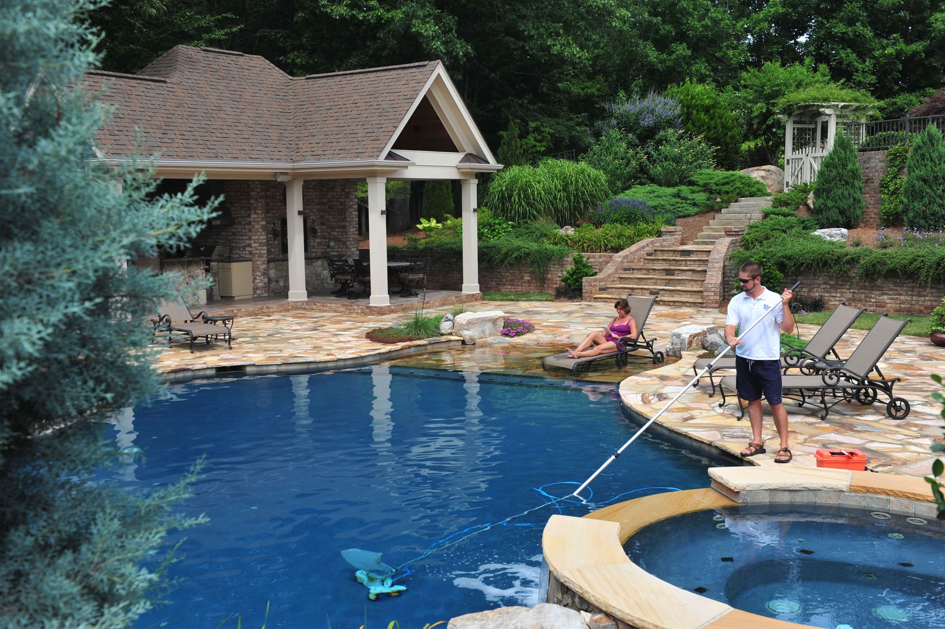 A person cleaning a backyard pool with a long-handled brush beside a stone patio