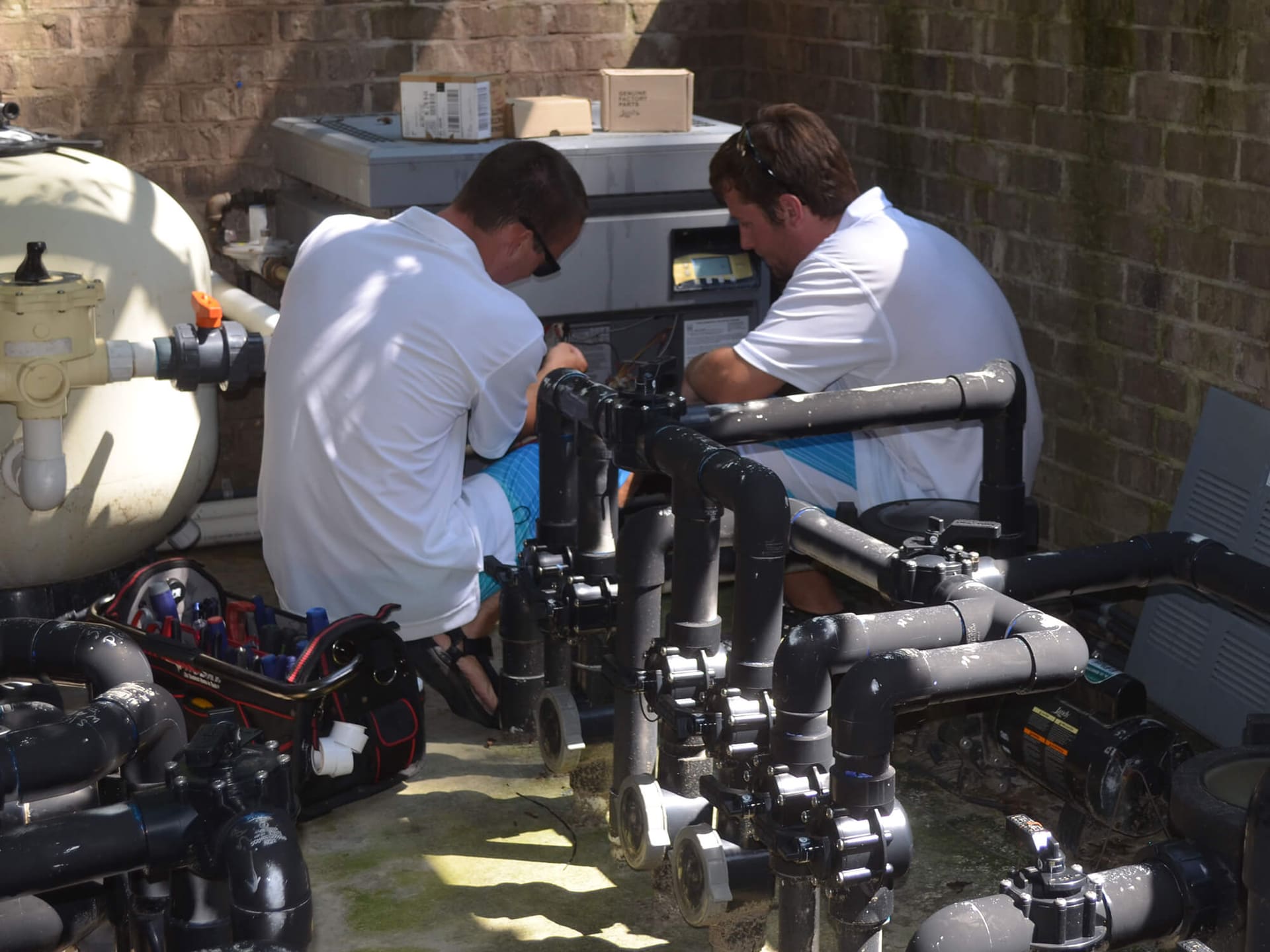 Two technicians working on a pool equipment pad with a filter, valves and plumbing